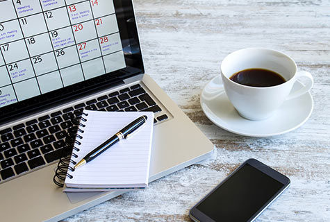 A laptop with a calendar on the screen next to a pen and notepad, cell phone, and cup of coffee